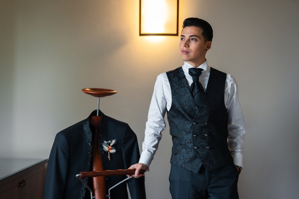 Groom is standing with one hand is his pocket, leaning with the other one on the coat rack where his wedding jacket is handing.