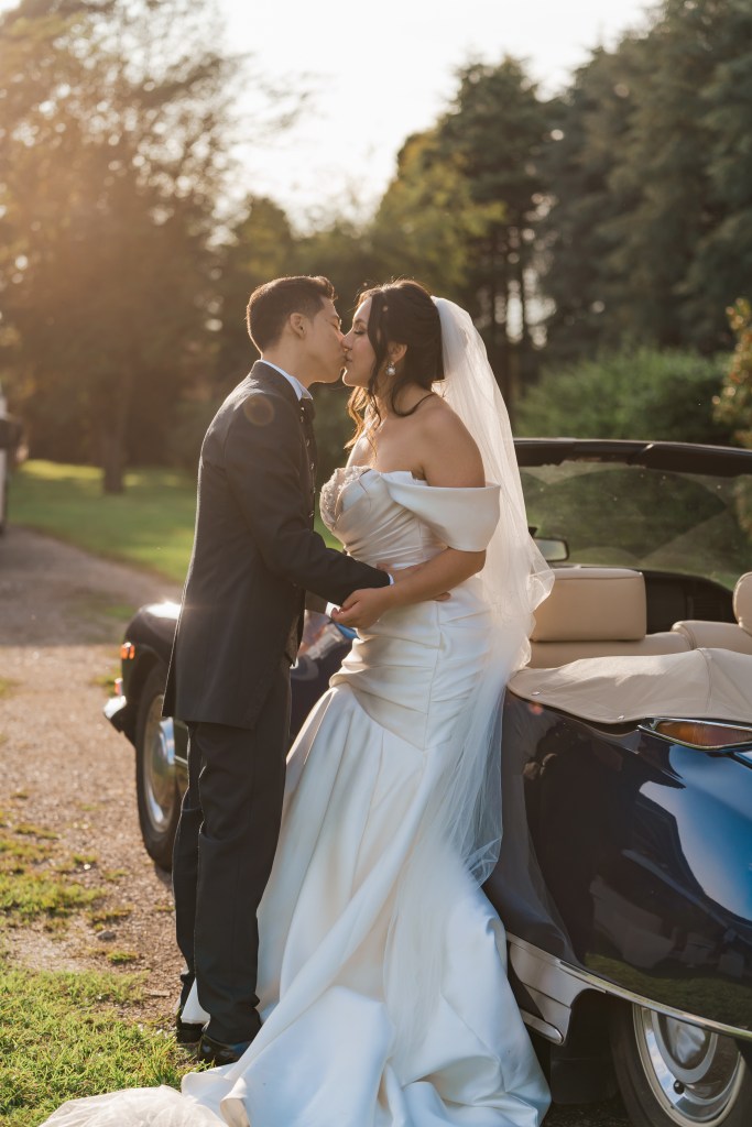 Bride and groom are kissing while leaning against a vintage car. It's sunset time and the sun gives a soft golden glare on the photo