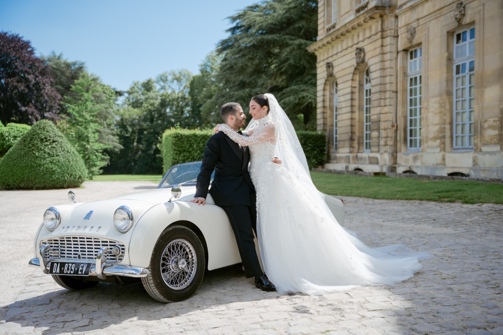 Bride and groom pose while leaning on a vintage while car next to Chateau de Champlareux