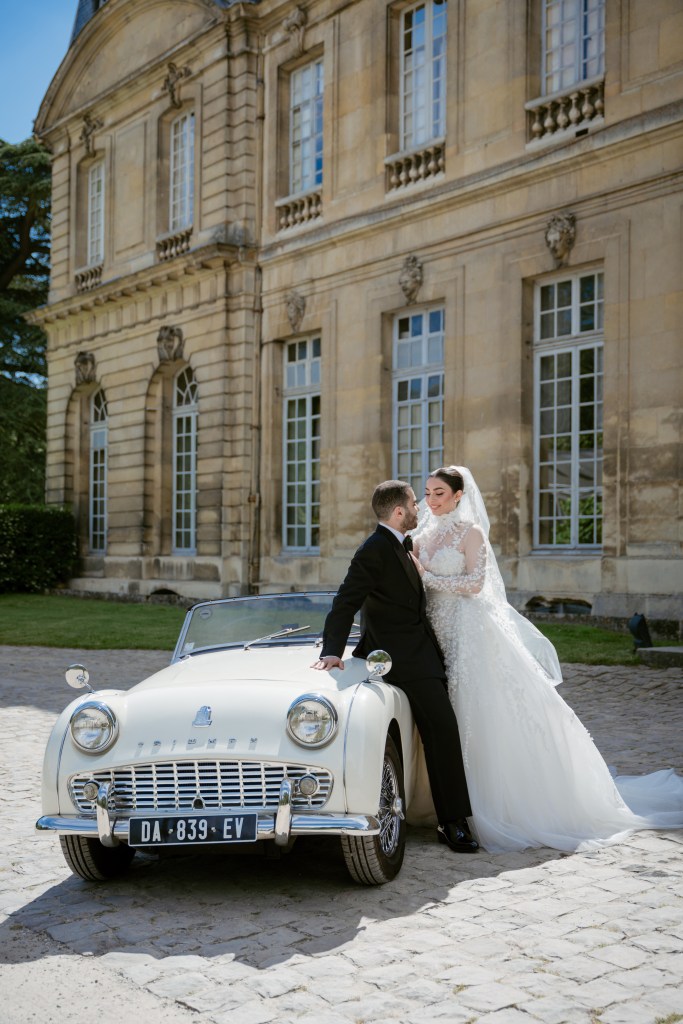 The groom is leaning on a white vintage car while holding the bride around her waist. They're posing in front of Chateau de Champlatreux