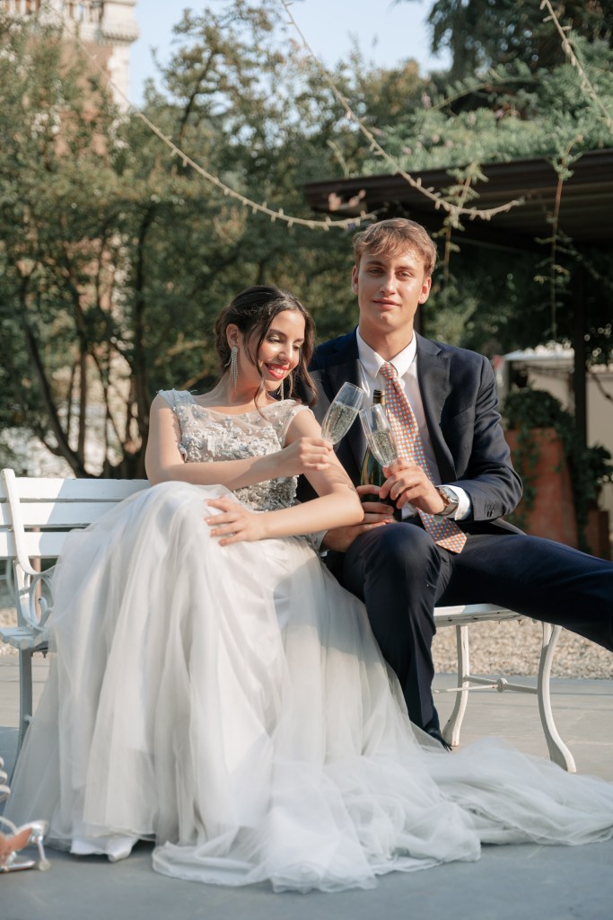 Bride and groom are sitting on a bench outside an Italian villa with champagne glasses in their hands. The bride is slightly leaning on the groom's shoulder