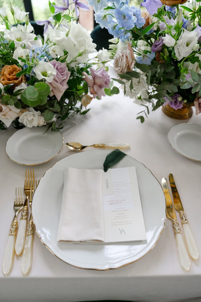 A close up shot of the dinner table decor with elegant golden cutlery and the dinner menu placed on the plate. In the middle are blue-purple-white flower arrangements