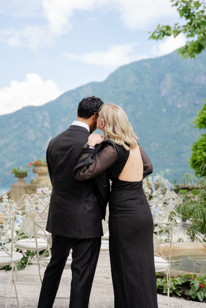 A quick candid shot of two elegantly dressed wedding attendees. They're turned with their backs towards the camera, the woman is whispering something into the man's ear