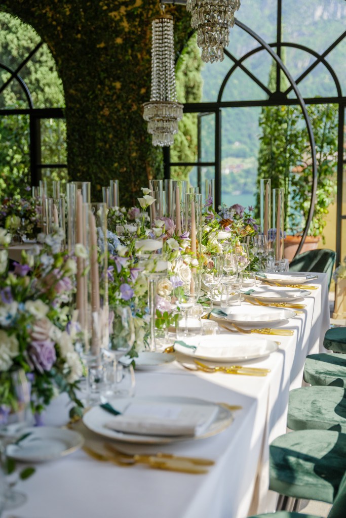 A close up shot of the wedding dinner decor. Everything is in green and yellow tones with a sprinkle of some purple flowers. There long candles on the table and decorative chandeliers above