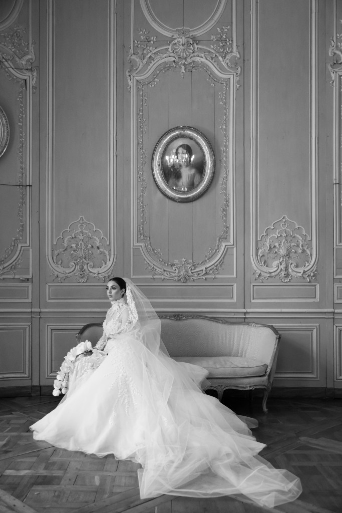 A black and white photo of a bride sitting on a sofa in the blue room of Chateau de Champlatreux. She's turned slightly sideways, the long dress train is laying nicely on the floor