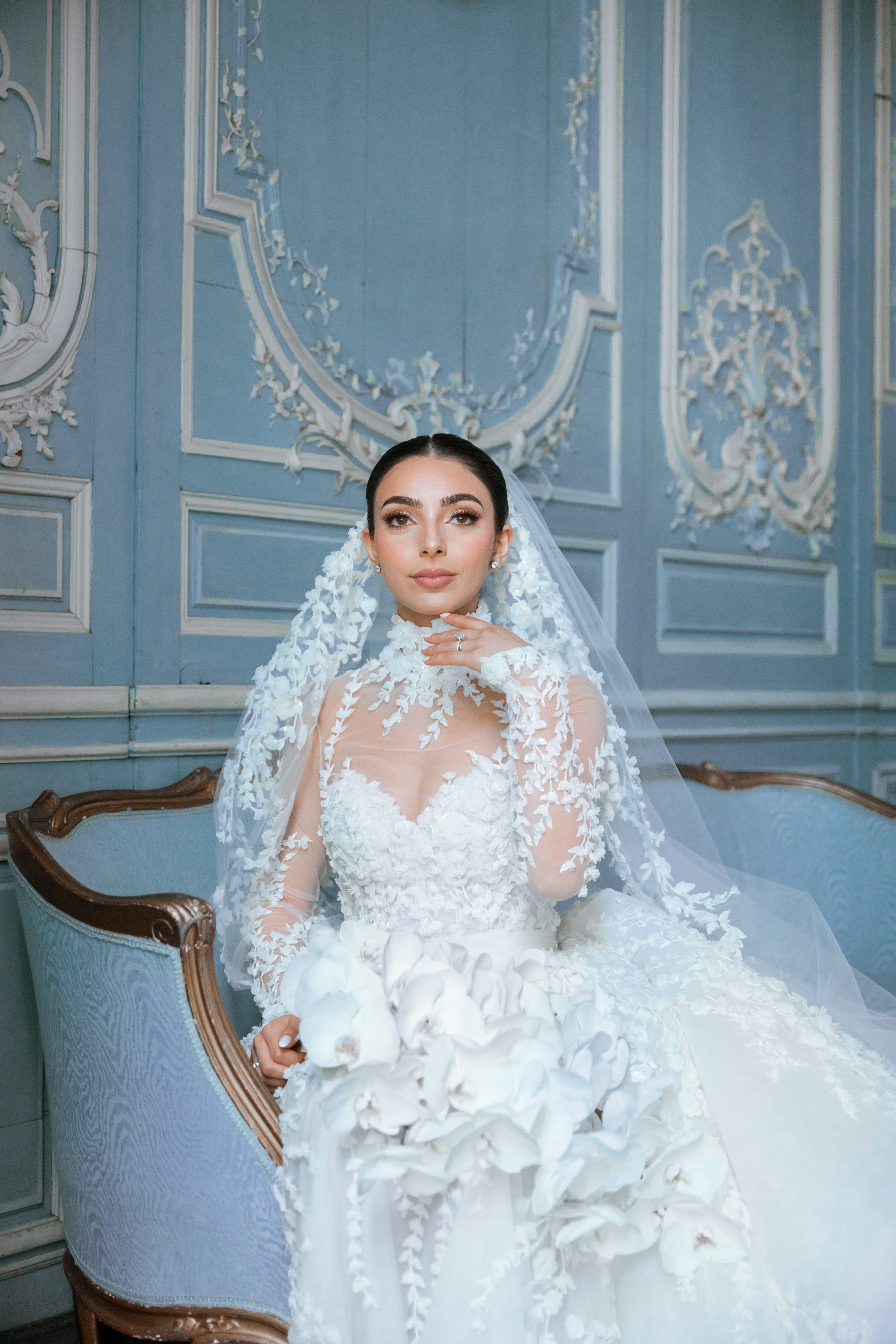 A soft portrait of a bride sitting on a sofa in the blue room of Chateu de Champlatreux. She's looking into the distance, with her hand gently placed under her chin