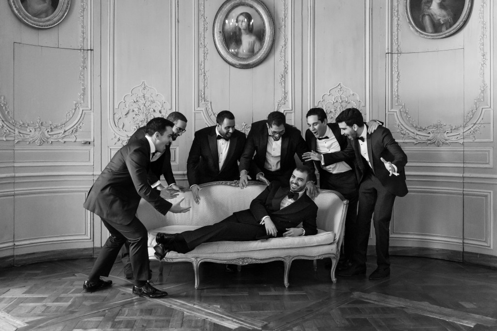 A black and white candid photo of the groom laying on a sofa in the blue room of Chateau de Champlatreux being surrounded in semi-circle by his groomsmen, all laughing