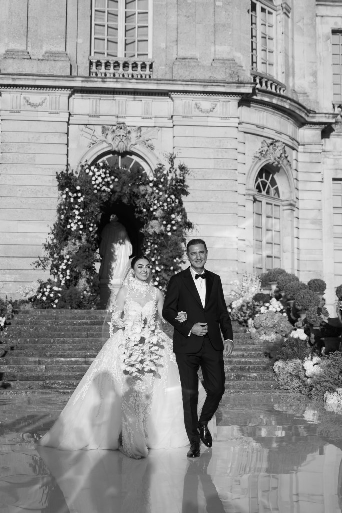 Bride is being led down the wedding isle by her father. Both are smiling. The photo is black and white