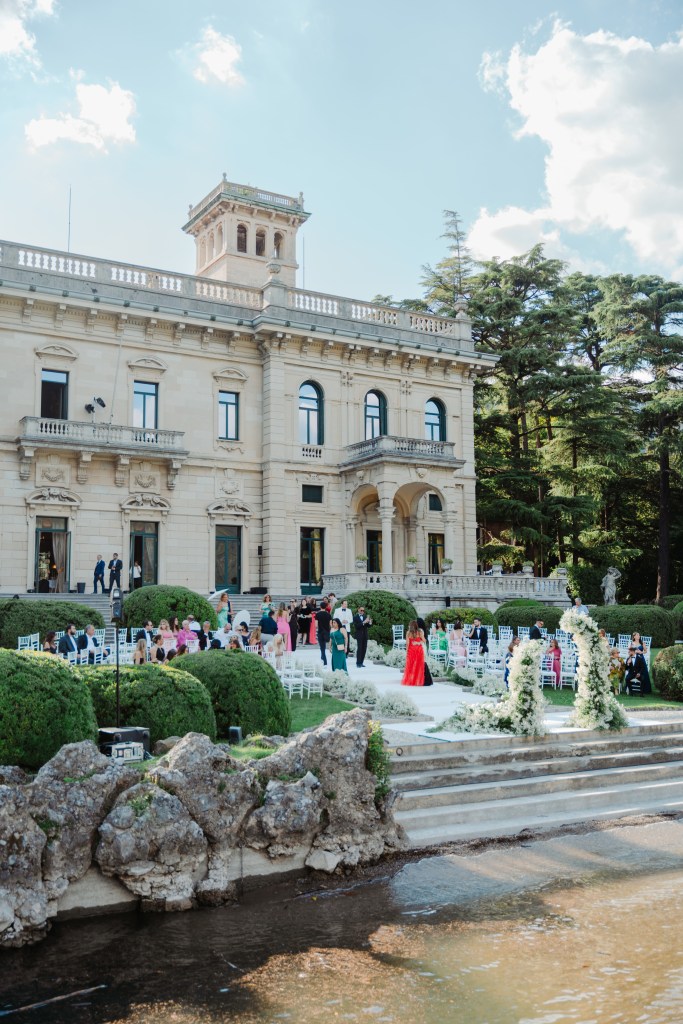 A far-out view of Villa Erba and the wedding ceremony set up in front of it, near the lakeside with guests mingling with each other