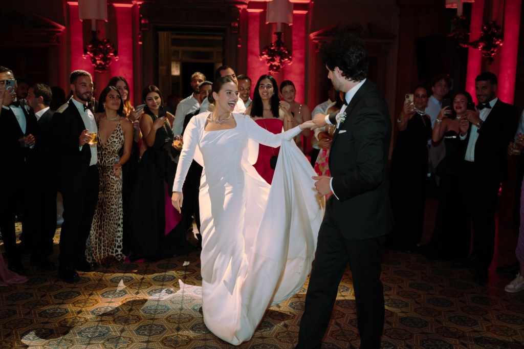 Newlyweds are in the middle of their first dance, surrounded by the wedding guests. The lights in the villa hall are very dimmed with the spotlight on the couple. The bride is looking back at the groom, smiling.