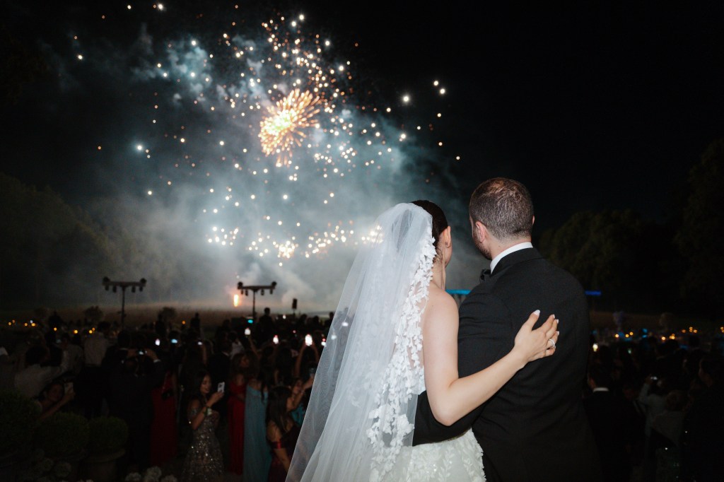 A shot of bride and groom standing turned away from the camera, hugging while watching the fireworks go off in the distance