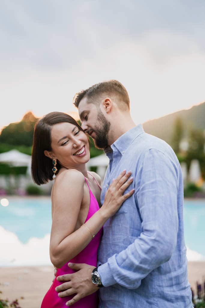 The couple is standing in the soft sunset light, holding each other. He is leaning in in kiss her on the cheeck, while she is turning her head towards the camera shyly smiling with her eyes closed