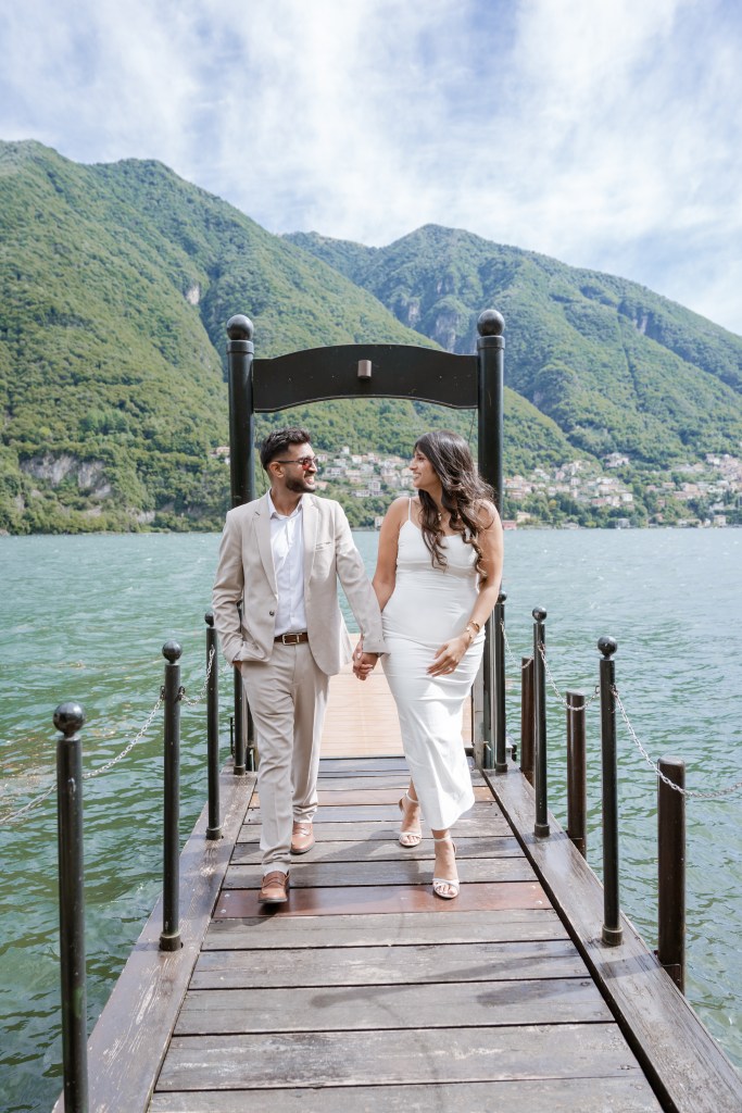 A couple dressed elegantly in white and beige colours is walking hand-in-hand on a small boat dock. In the bacground is Lake Como with its green mountains and blue water