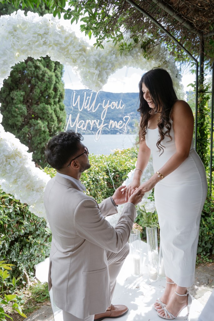Shot of a suprise proposal at Lake Como. The couple is standing next to a heart-shaped arch with "will you marry me" neon sign in the middle. He is standing on one knee, she is shocked and happy