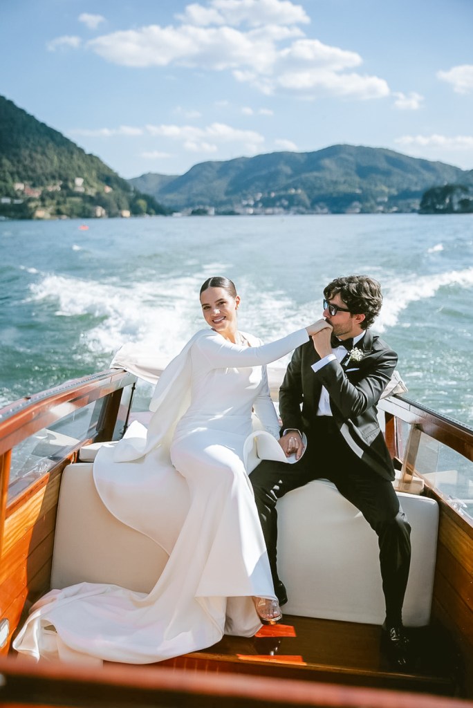 The newlyweds are sitting at the back of a wooden boat sailing across Lake Como. The groom is gently kissing the bride's hand
