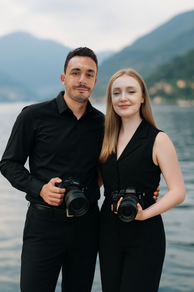 Alber and Anastasia are Lake Como wedding photographers and videographers. They are standing together, dressed in all black, holding their Sony cameras and smiling into the camera. Lake Como and its mountains are in the background