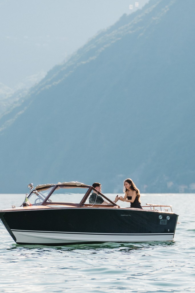 A couple is in a wooden boat in the middle of Lake Como. The man is kneeling to propose, the woman is happily surprised