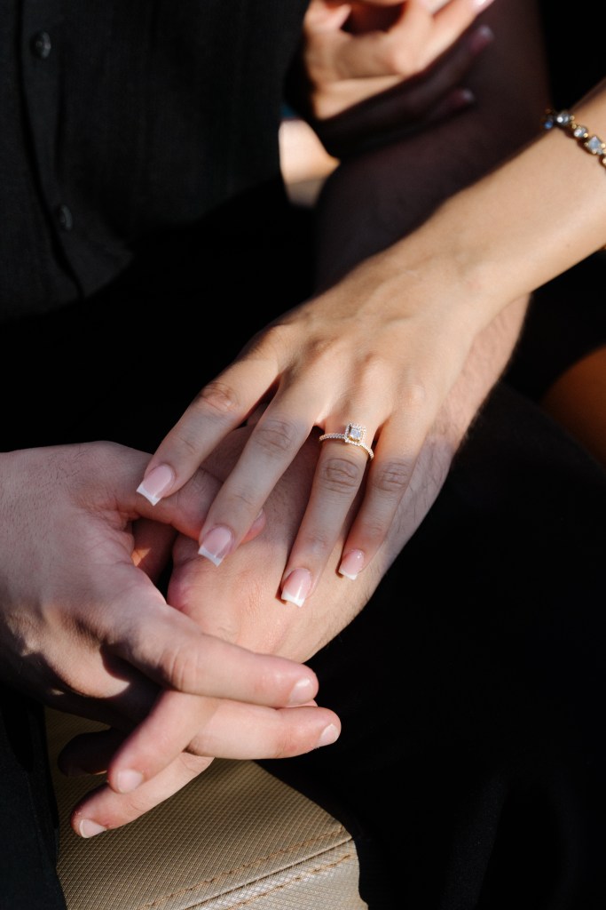 A close up photo of the couple's hands intertwined on their knees. The focus of the photo is the new engagement ring